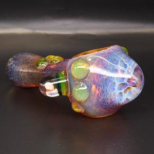 May include: A hand pipe made of iridescent glass featuring a swirling pattern of purple, blue, and orange hues. The bowl area is rounded and translucent, accented with small, raised green glass dots along the side and a clear glass mouthpiece.