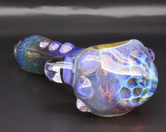 May include: A hand pipe made of iridescent glass featuring a deep blue and purple color palette with a honeycomb pattern on the bowl. The stem has a textured surface with small raised glass bumps, and the overall finish has a metallic sheen.