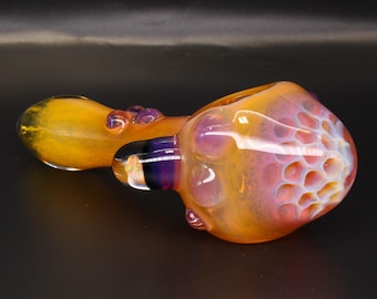 May include: A hand pipe made of glass featuring a yellow stem and a bowl with a honeycomb pattern in shades of orange, pink, and purple. The glass has a smooth, glossy finish with a rounded shape and a small dark purple accent near the bowl.