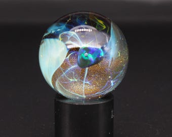 May include: A spherical glass paperweight featuring swirling patterns of blue, teal, and white glass with embedded gold-toned glitter particles. The orb is mounted on a solid black cylindrical base, creating a contrast between the clear glass and the dark stand.