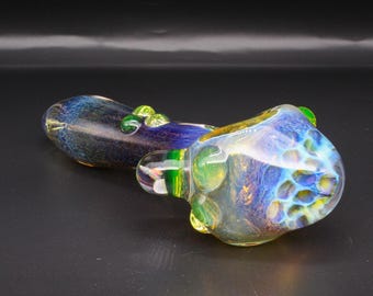 May include: A hand pipe crafted from glass featuring a deep blue and purple gradient body with a textured, honeycomb-like pattern on the bowl. The piece includes translucent green glass accents and a clear glass mouthpiece, showcasing a smooth finish.