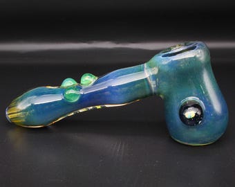 May include: A hand pipe crafted from translucent glass featuring a deep blue and teal gradient color scheme. The pipe includes a bowl section with a dark circular accent and a stem adorned with two small, raised green glass beads on the top surface.