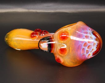 May include: A hand pipe made of glass featuring a gradient of orange and yellow hues. The bowl section displays a textured honeycomb pattern with iridescent purple and pink highlights, accented by small, raised red glass dots along the stem.