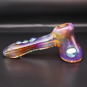 May include: A glass hand pipe featuring a hammer-style design with an iridescent finish that shifts between shades of purple, gold, and orange. The pipe includes a circular bowl, a long stem with three small glass beads, and a side carb hole.