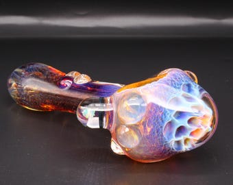 May include: A hand-blown glass pipe featuring a deep amber and purple stem with a swirling pattern. The bowl end displays an iridescent, multi-colored finish with a textured, honeycomb-like structure and several clear, rounded glass protrusions.