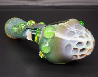 May include: A glass pipe featuring a multi-colored iridescent finish with a honeycomb-textured bowl. The stem is adorned with several raised, bright green glass beads and a central band of dark green and clear glass accents along the side.