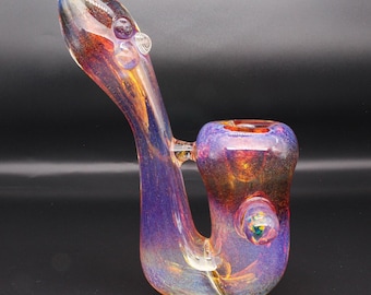 May include: A glass pipe featuring an iridescent, color-changing finish that shifts between shades of purple, blue, and amber. The piece has a curved, elongated neck, a rounded bowl section, and a small circular carb hole on the side of the bowl.