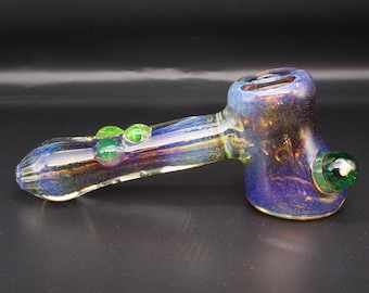 May include: A glass hand pipe featuring a deep blue and iridescent gold finish. The pipe has a cylindrical stem with two small green glass beads attached to the top and a larger green glass bead on the side of the bowl, which has a wide opening.