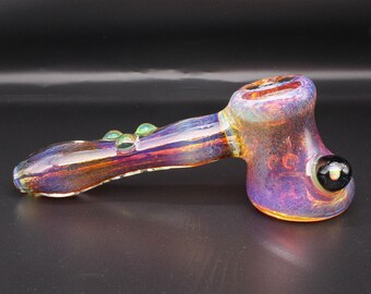 May include: A hand pipe made of iridescent glass with a metallic sheen, featuring a bowl with a black glass marble accent on the side and three small green glass dots along the stem, showcasing purple, gold, and blue color shifts throughout.
