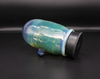 May include: A glass vessel featuring a rounded, iridescent body with swirling shades of teal, blue, and green. The piece has a smooth, glossy finish, a small protruding blue glass knob on the side, and a flat, circular black base at the opening.