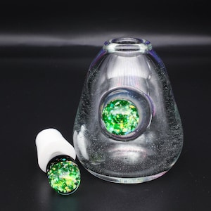 May include: Clear glass bottle with a white stopper. The bottle has a large, round, green and yellow speckled design in the center. The stopper has a matching design. The bottle is on a black surface.