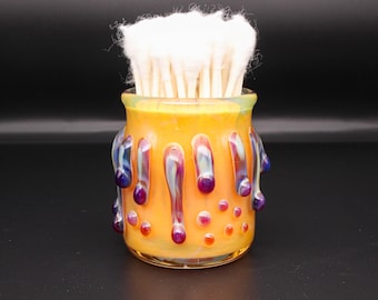 May include: A cylindrical glass holder with a vibrant orange base, decorated with iridescent purple and blue drip-like accents and small raised dots. The container is filled with a cluster of white cotton swabs with wooden stems.
