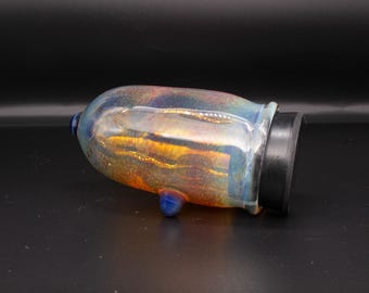 May include: A cylindrical glass vessel with an iridescent finish displaying shifting hues of blue, gold, and amber. The object features a small blue glass protrusion on its side and is capped with a solid black circular lid at one end.