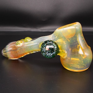 May include: A hand pipe made of iridescent glass with a yellow and orange hue. The pipe features a prominent dark green circular carb cap with embedded glitter and two small yellow glass decorative accents on the stem near the mouthpiece.