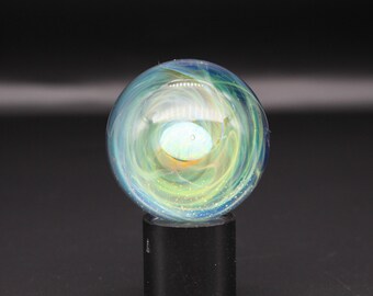 May include: A spherical glass marble featuring swirling patterns of blue, green, and white colours with a central iridescent orb. The marble is clear and contains small reflective flecks throughout, resting on a solid black cylindrical stand.