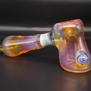 May include: A colorful glass pipe with a hammer-like shape. The pipe features a blend of orange, yellow, and purple hues with a blue swirl design. The glass has a speckled appearance and a white band near the mouthpiece.