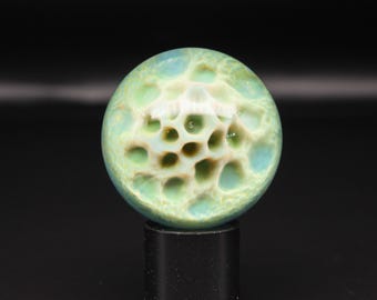 May include: A spherical marble featuring a translucent light blue and green surface with a textured, cream-coloured honeycomb pattern of recessed circular indentations across the centre, resting on a small black cylindrical stand.