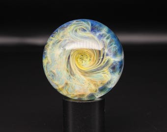 May include: A spherical glass marble featuring a swirling internal pattern of yellow, white, and blue colours. The glass orb has a smooth, polished surface with a reflective finish, showcasing intricate, cloud-like wisps of colour throughout the sphere.