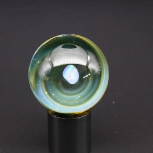 May include: A handcrafted glass marble with a swirling design of green, yellow, and blue hues. The marble features an internal, iridescent, oval-shaped inclusion. It is displayed on a black stand, showcasing its unique artistry.