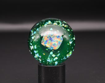 May include: A spherical glass paperweight in a deep forest green hue, featuring a central iridescent rectangular inlay with a rainbow colour shift. The sphere is filled with numerous small, reflective, light-catching flakes in shades of blue, green, and silver.