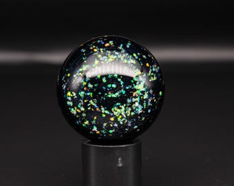 May include: A spherical object with a glossy black base featuring embedded iridescent flakes in shades of green, yellow, and blue. The sphere is mounted on a cylindrical black stand, creating a contrast between the dark background and the light-reflecting particles scattered throughout the surface of the round item.