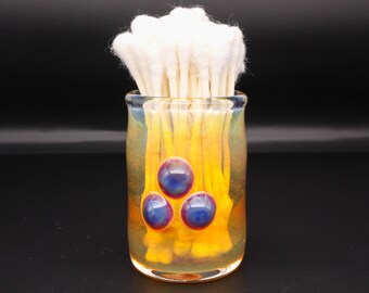 May include: A cylindrical glass holder with a translucent yellow and iridescent finish, featuring three embedded blue and purple circular glass cabochons on the front. The holder is filled with a cluster of white cotton swabs with paper stems.