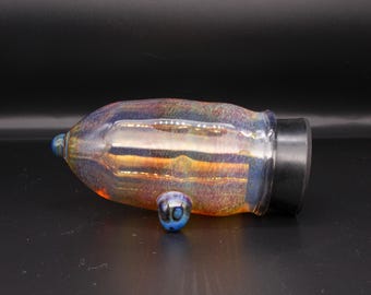 May include: A cylindrical glass vessel with an iridescent, multi-coloured finish featuring hues of orange, blue, and gold. The object has a rounded, tapered end with a small protrusion, a single blue glass foot, and a solid black circular base cap.