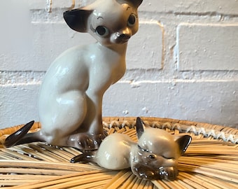 May include: Two ceramic Siamese cat figurines. The larger cat is sitting up with its tail curled around its legs. The smaller cat is lying down with its head resting on its paws. Both cats are painted in shades of white and gray.