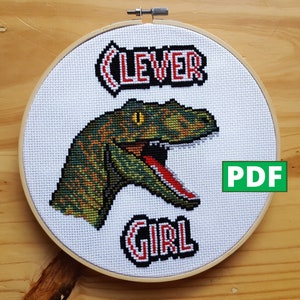 Può includere: Ricamo a punto croce in un cerchio di legno, raffigurante un dinosauro verde, marrone e giallo con la bocca aperta. Il testo "CLEVER GIRL" è ricamato in rosso e nero. Un'icona "PDF" verde è visibile.