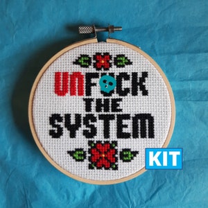 May include: Cross-stitch embroidery hoop with the phrase "UNFUCK THE SYSTEM" in red and black. The design includes floral accents and a teal skull. The word "KIT" is in a blue box. The embroidery is set in a wooden hoop.