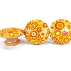 May include: Three decorative wooden knobs with a retro floral design in shades of yellow, orange, and pink. One knob is mushroom-shaped, while the other two are round. The knobs have a natural wood base.