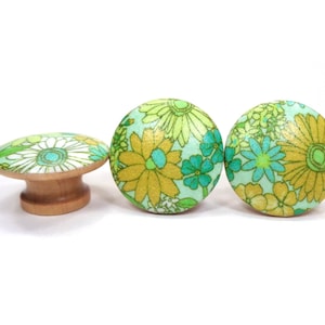 May include: Three wooden cabinet knobs with a floral design. The knobs feature a light green background with yellow, green, and blue flowers. One knob is a flat oval shape, and the other two are round.