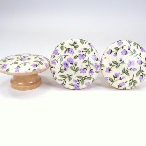 May include: Three wooden knobs with a white background and a purple floral pattern. The knobs are round and have a small stem on the back.