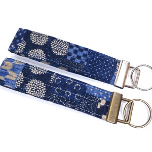 May include: Two blue and gold patterned fabric keychains with metal key rings. The keychains are made of a patchwork fabric with a floral design.