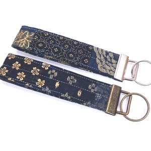 May include: Two fabric wrist keychains with floral and geometric patterns in navy blue and gold. One keychain has a silver-tone metal ring, and the other has a bronze-tone metal ring.