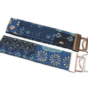May include: Two blue and gold patterned fabric keychains with metal key rings. The keychains are made of a patchwork of different fabrics with gold accents.