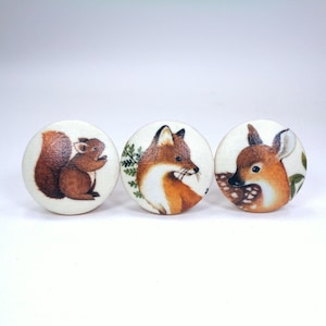 May include: Three round wooden cabinet knobs with woodland animal illustrations. The knobs feature a squirrel, a fox, and a fawn. The squirrel is brown, the fox is orange and white, and the fawn is brown and white.