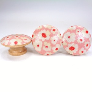 May include: Three pink and white floral patterned wooden knobs. The knobs have a round top and a short, cylindrical base.