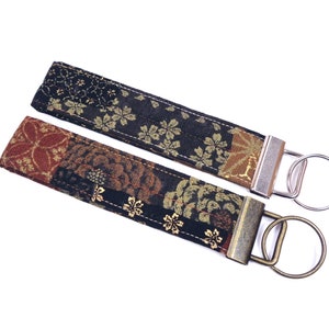 May include: Two fabric keychains with floral patterns. One keychain is black with gold and brown flowers, the other is brown with gold and red flowers. Both keychains have metal rings.