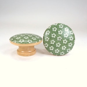May include: Two green and white floral patterned wooden knobs. The knobs have a round top and a short, cylindrical base.