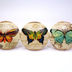 May include: Three round wooden knobs with a vintage design featuring different colored butterflies. The butterflies are printed on a cream background with a faded script.
