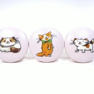 May include: Three round, light pink cabinet knobs with hand-drawn cat illustrations. One cat is brown and white, another is orange with a green bow tie, and the third is white and brown. These knobs are perfect for a child's room or a cat lover's home.