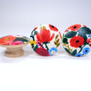May include: Three decorative wooden knobs with floral designs. Two are round, featuring red poppies, blue and yellow flowers on a white background. The third knob is mushroom-shaped, with a similar floral pattern. The knobs have a natural wood base.
