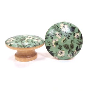 May include: Two wooden knobs with a green floral pattern. The knobs are round and have a small stem.