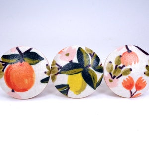 May include: Three round fabric buttons with a white background and a floral pattern featuring oranges, lemons, and pink flowers.