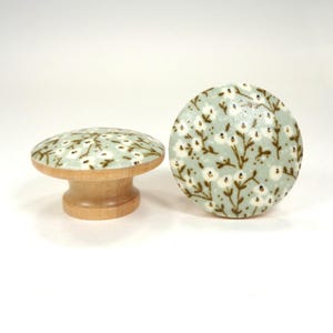 May include: Two decorative wooden cabinet knobs. The knobs have a light blue background with a white floral pattern and brown stems. One knob is round, and the other is mounted on a wooden base.