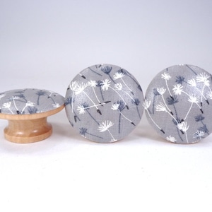 May include: Three decorative cabinet knobs. Two are round, and one is a mushroom shape. They feature a gray fabric with a white and blue floral pattern. The bases are made of light-colored wood.