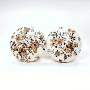 May include: Two round, white cabinet knobs with a floral design. The design features brown and tan flowers and leaves on a white background. The knobs are set against a plain white background.