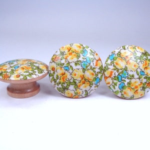 May include: Three floral cabinet knobs. Two are round, and one is a mushroom shape. The knobs feature a white background with yellow, orange, and blue floral patterns. The bases are made of light-colored wood.
