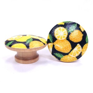 May include: Two wooden cabinet knobs with a lemon design. The knobs have a light wood base and a round top with a black background and yellow lemons and green leaves. One knob is round, and the other is a mushroom shape.